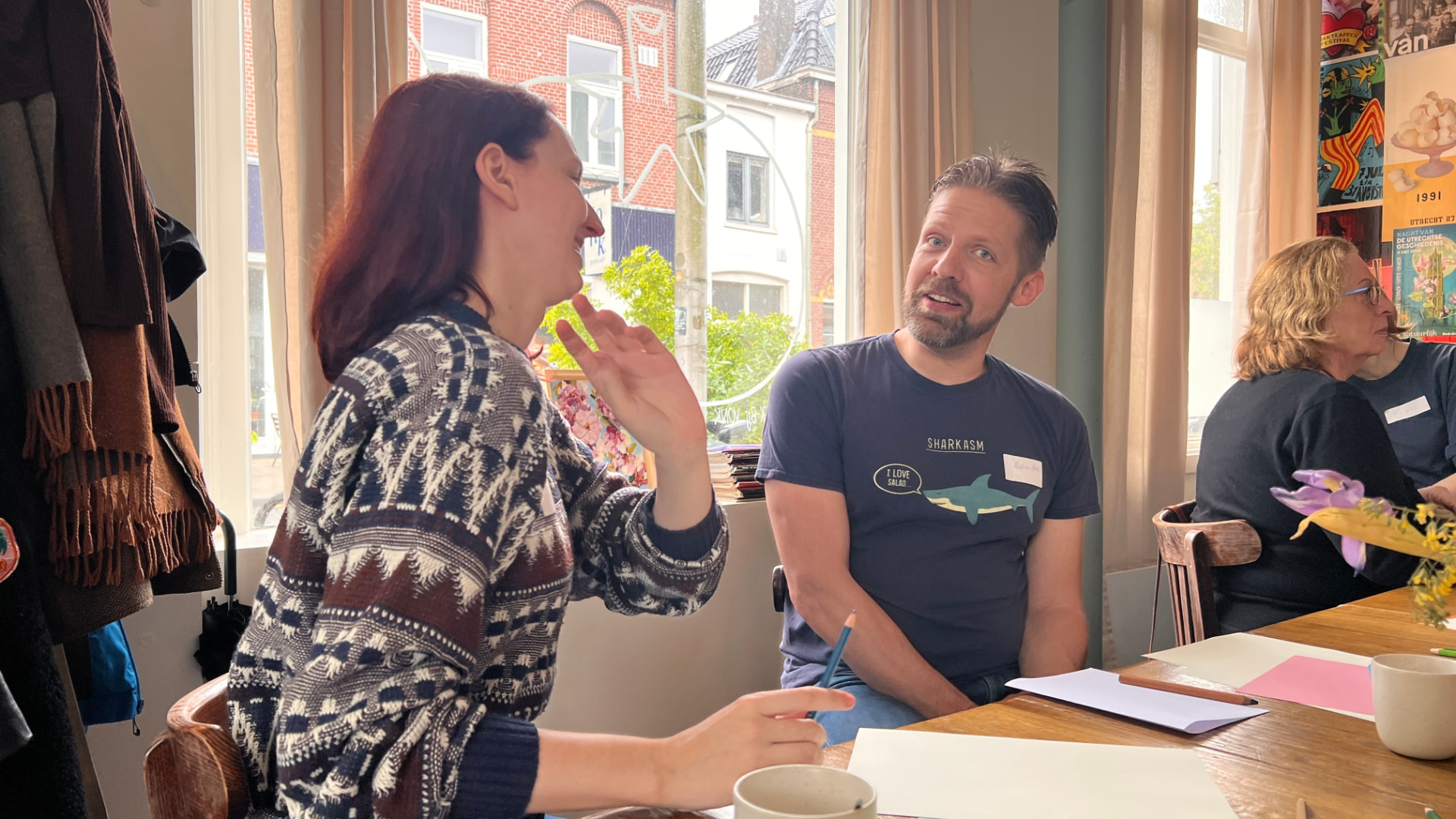Een vrouw en een man met naamstickers praten lachend met elkaar aan een houten tafel bij een groot raam, met papier en koffiekopjes voor zich