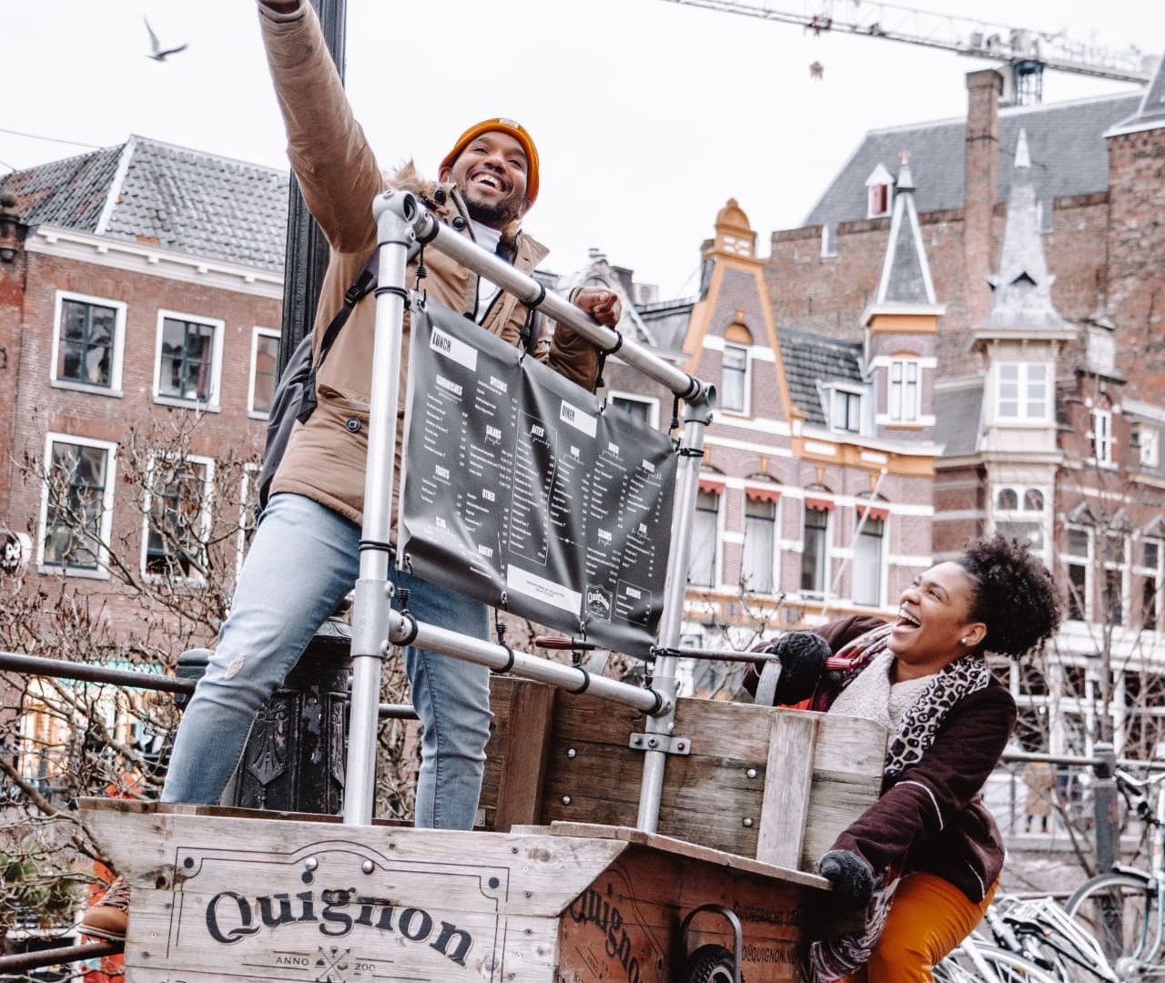 Twee lachende mensen bij een houten bakfiets op een Utrechtse kade: een man staat in de bak en wijst omhoog, een vrouw duwt de kar voort langs bakstenen gevels