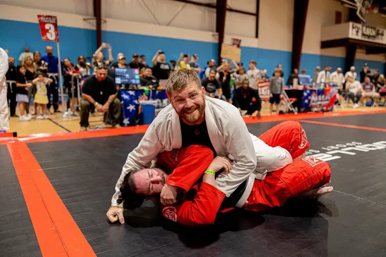 Twee mannen trainen Braziliaans Jiu-Jitsu op een zwarte mat met oranje belijning; de man bovenop in een witte jas en rode broek lacht naar de camera terwijl zijn tegenstander in een rood pak onder hem ligt, met toeschouwers en wedstrijdtafels op de achtergrond
