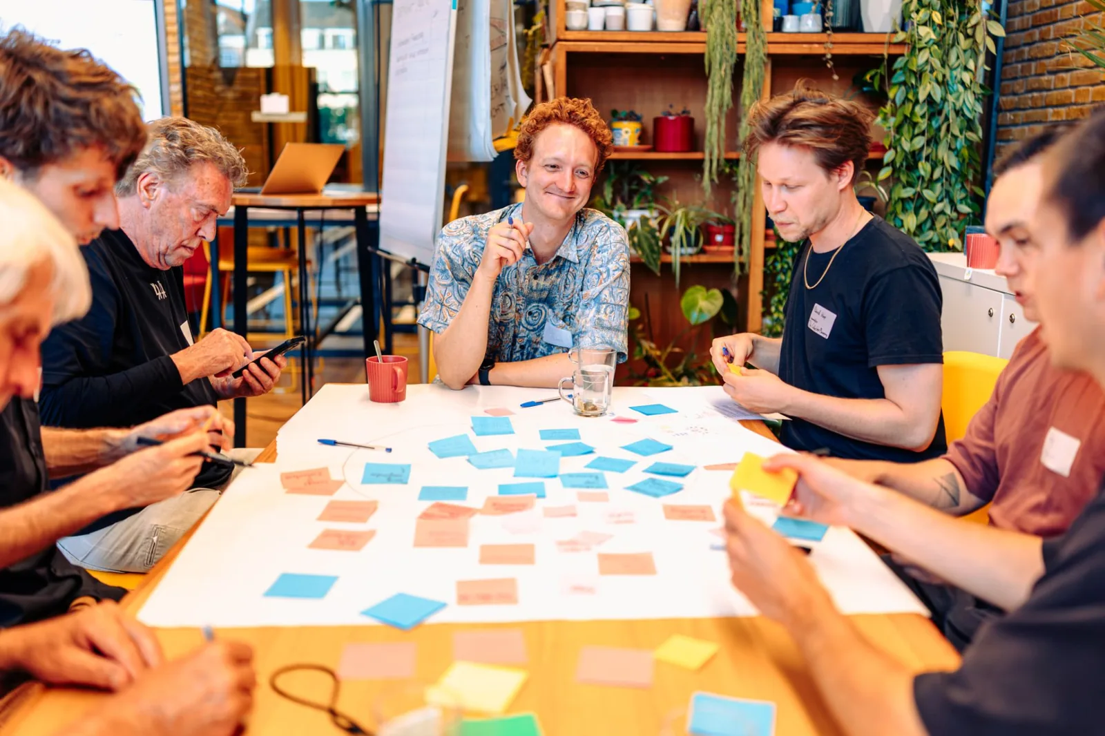 Deelnemers van de stuurgroep Coöperatieve Economie zitten aan een tafel met naamstickers en papieren, met een flapover met gekleurd papier op de achtergrond