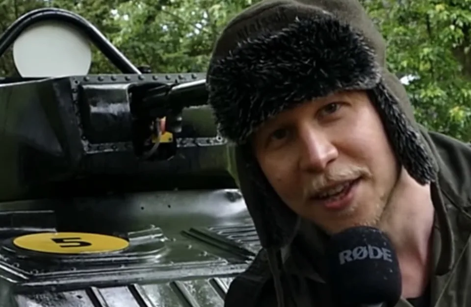 Een man in een legergroene jas en bontmuts spreekt in een microfoon richting de camera, met een oud pantservoertuig op de achtergrond tussen de bomen